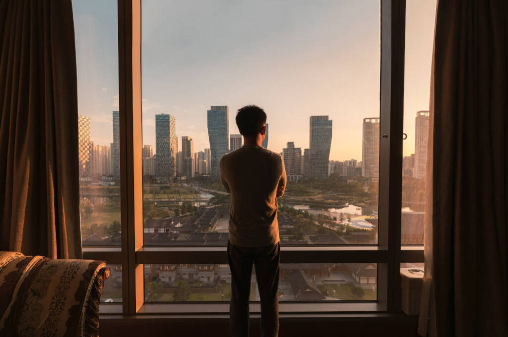 Young asian man standing and looking through window in a hotel. Sunrise over modern buildings in Songdo central park at Incheon, South Korea