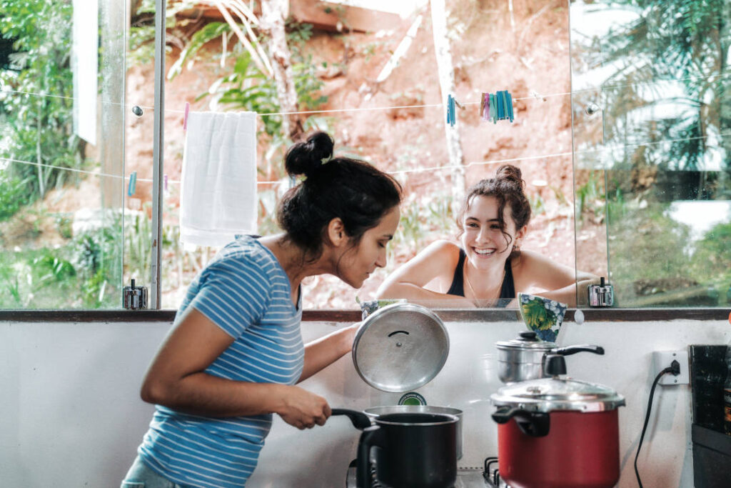 side view on woman lifting lid to check the meal on stove while smiling daughter watching scene from outdoors through kitchen window