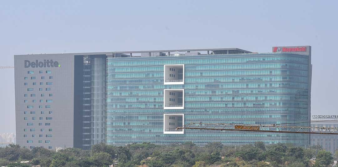 It is a representation of a modern glass office complex with two main buildings with green reflective facades. These imposing structures have numerous windows and display geometric and angular architecture. An entrance portico links the two buildings, emphasising the main entrance. Surrounding the building is a landscaped area with trees and green spaces, as well as a road with a car, evoking a planned and spacious urban environment.