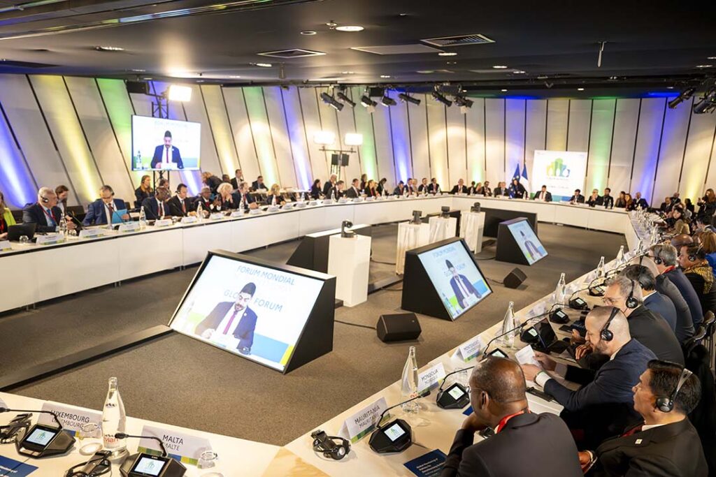 A photo of an international conference in progress. Delegates from various countries are seated around a large oval table, concentrating on the presenter's speech, which is broadcast on large screens around the room. Plaques with the names of the countries can be seen, as well as headphones for translation. The room has a modern feel, with soft lighting and neutral colors.